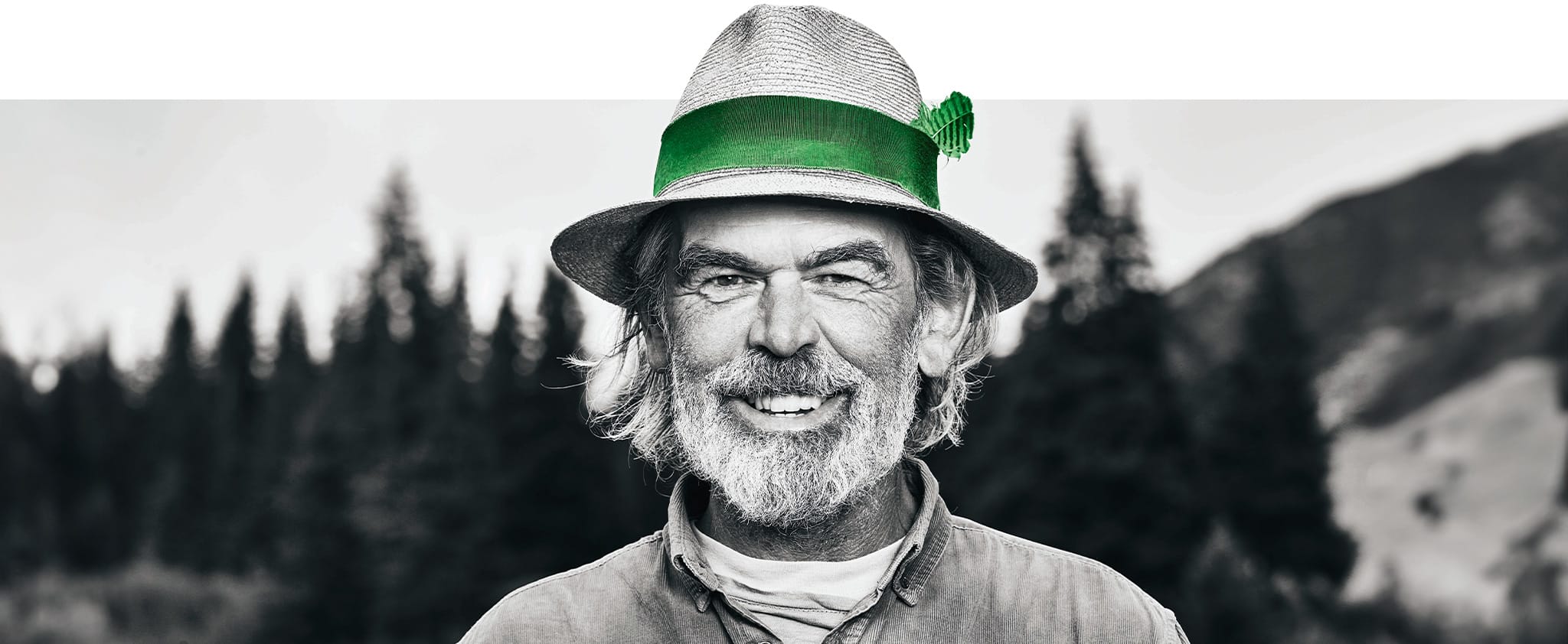 Ein älterer Mann mit grauen Haaren und Bart trägt einen Hut mit einem grünen Band und lächelt in die Kamera, mit einem verschwommenen Wald im Hintergrund.